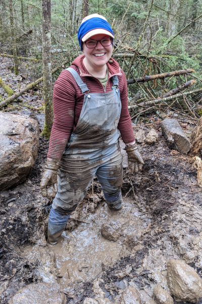 An Alliance volunteer smiles as she stands shin deep in mud.