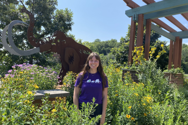 A young woman stands among wildflowers in front of a metal mammoth statue on a sunny summer day.