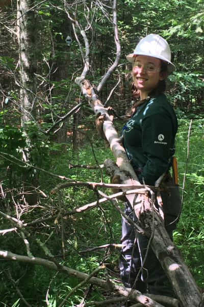 A WisCorps crew member in protective gear smiles as she holds a large tree branch.