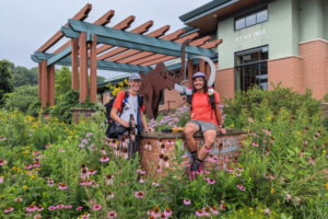 Two men on a long distance hike pose and smile together next to a metal mammoth statue with their hiking gear.