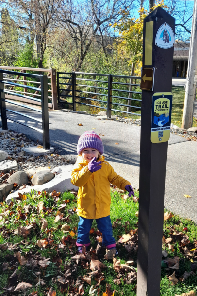 A kid in a yellow coat points to an Ice Age Trail post with a yellow blaze and Ice Age Trail Community sign.