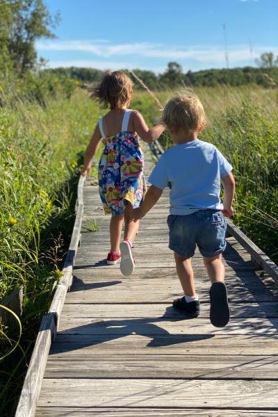 Two kids run across a boardwalk in a wetland on a sunny day.