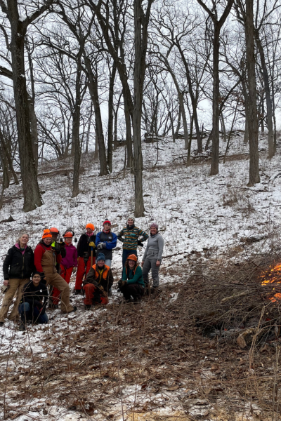 A group of smiling volunteers pose next to a burning brush pile.