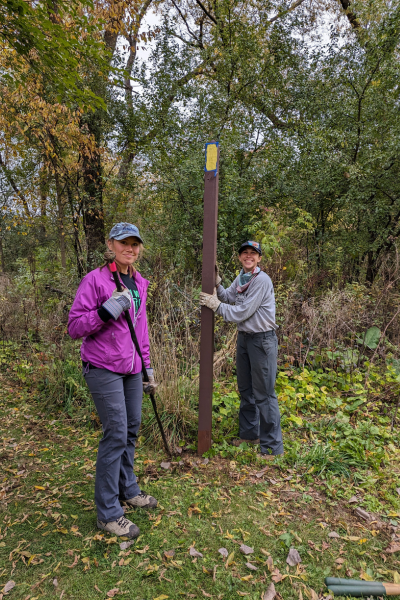 Two volunteers work to install a wooden post with a painted yellow blaze in the ground.
