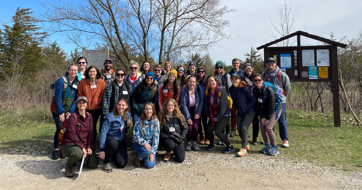 A group of young adults smile and pose together in front of a trailhead on an early spring day before a hike.